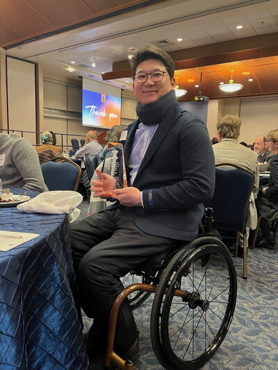 A man in a suit with glasses in a wheelchair holding a teaching award.