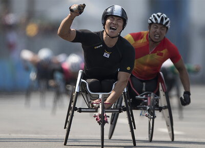A man in a racing wheelchair and a helmet clenches his fist in victory as another man in a wheelchair behind him races to catch him.